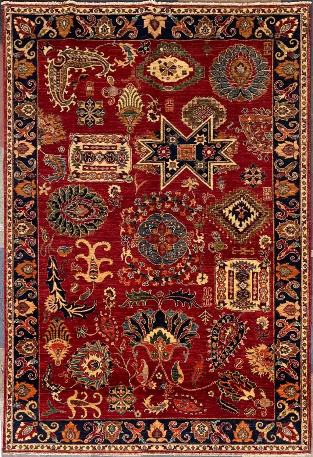 crimson-red-tribal-sampler-chobi-peshawar-wool-area-rug-6x9.webp | McFarlands Carpet and Rug Service Hand-knotted Chobi Peshawar wool area rug 6.8' x 9.2' with crimson red field, geometric star sampler motifs, and a navy blue border.