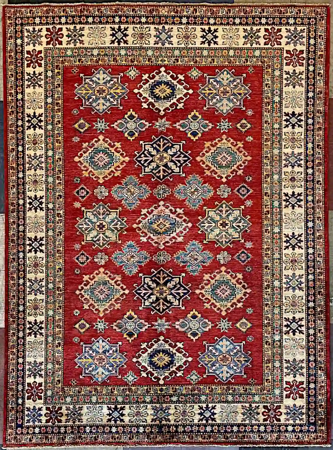 traditional-crimson-cream-kazak-wool-area-rug-6x9.webp | McFarlands Carpet and Rug Service Traditional crimson Kazak hand-knotted wool area rug 5.9' x 7.8' featuring a central row of navy star shields and a wide cream tribal border frame.